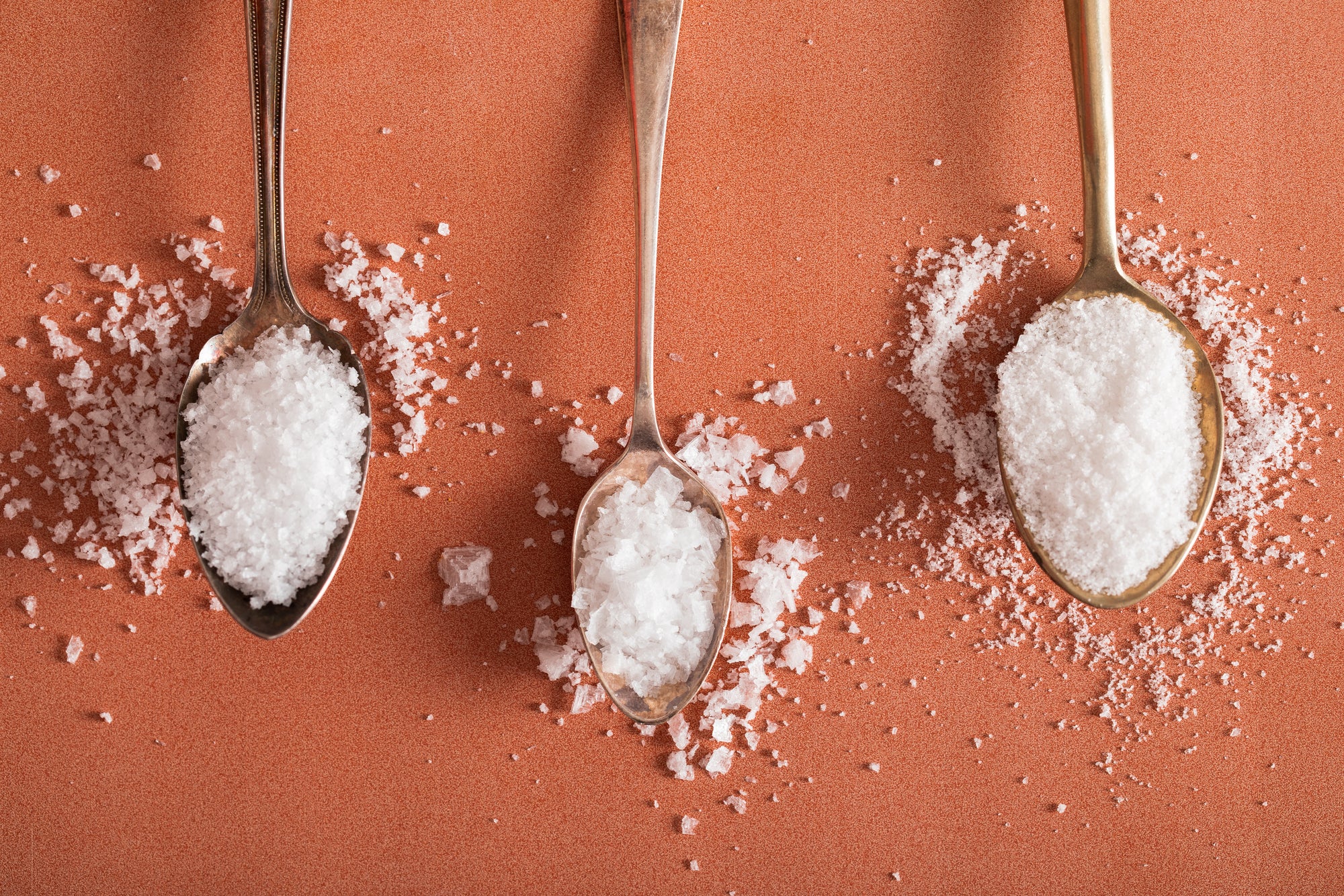 Flake, Kosher and Fine sea salts each on a spoon on an orange tabletop.