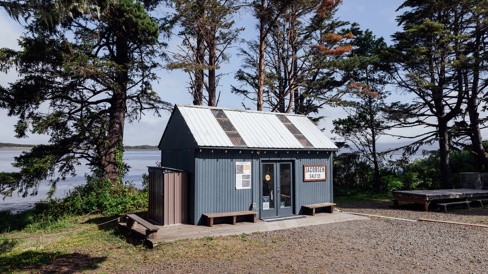 A small blue house structure next to the waters of Netarts Bay surrounded by sparse trees. The Jacobsen Salt Co gift shop.