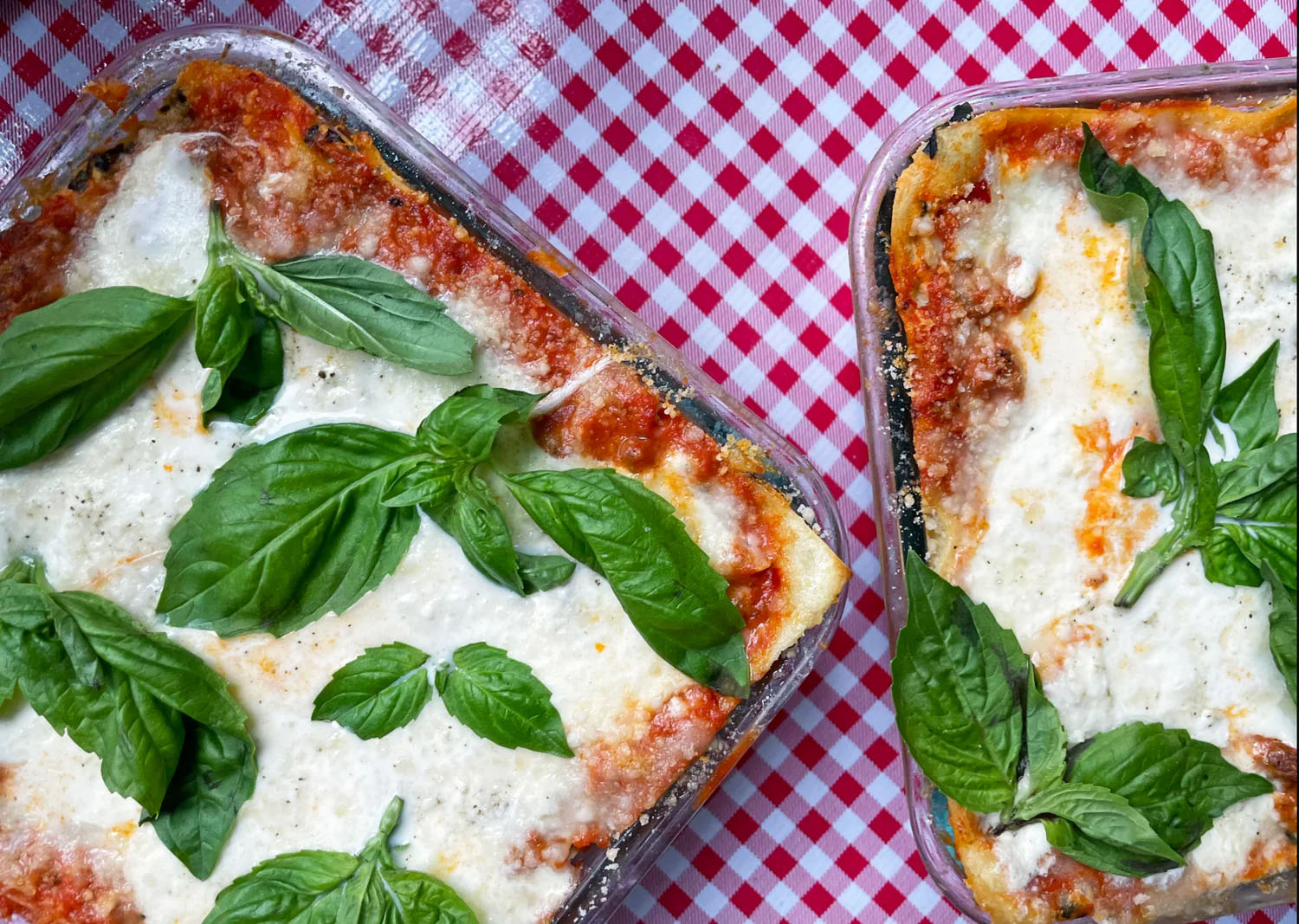 Two trays of freshly baked lasagna sit on top of a red gingham tablecloth.