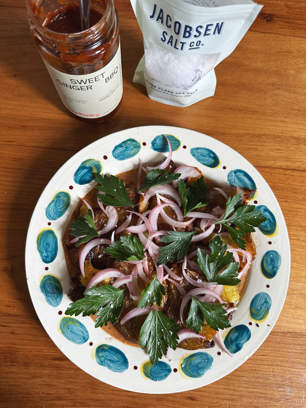 Crispy potatoes, plated with pickled onions and parsley. Jacobsen salt and a jar of BBQ sauce sit above the plate.