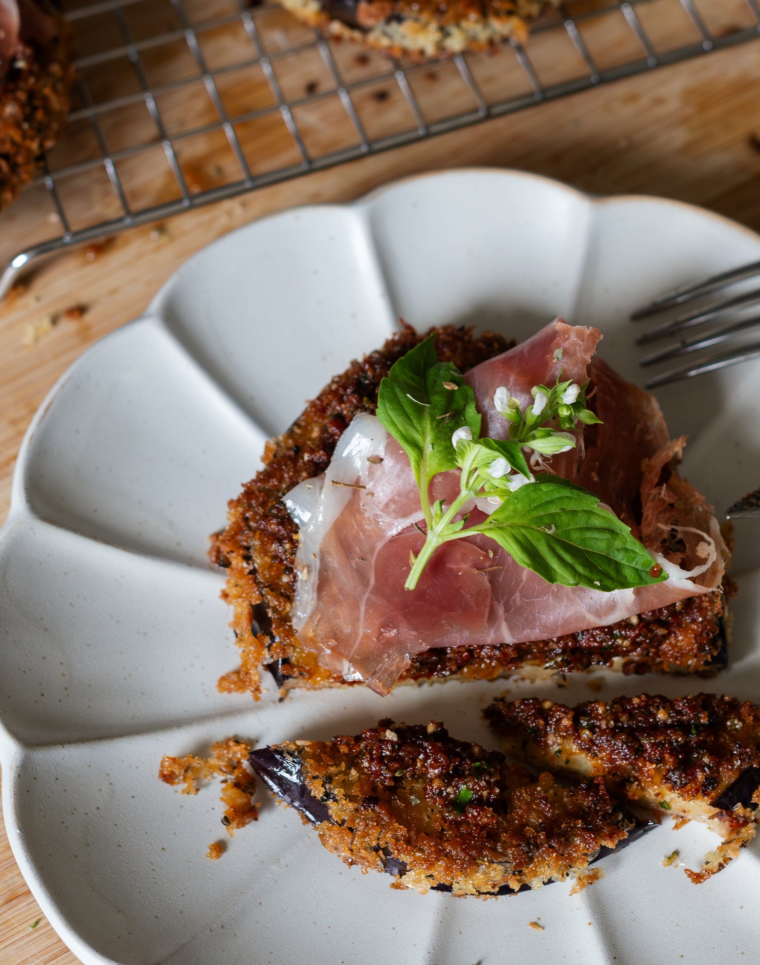 Breaded eggplant on a plate, topped with prosciutto and a sprig of basil.