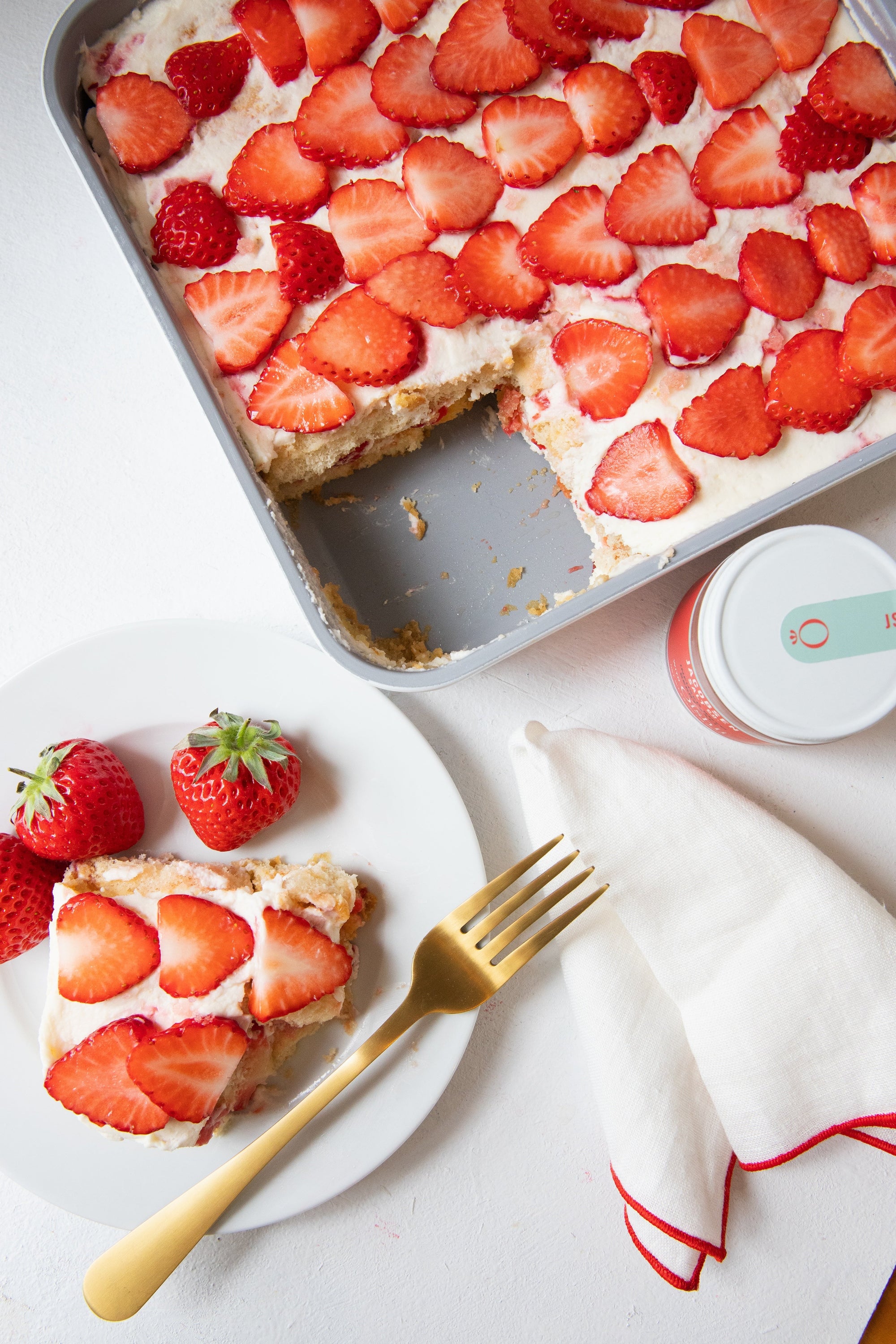 White cake topped with sliced strawberries, with a square sliced out of the corner. The sliced piece of cake appears at the bottom left, plated with whole strawberries.