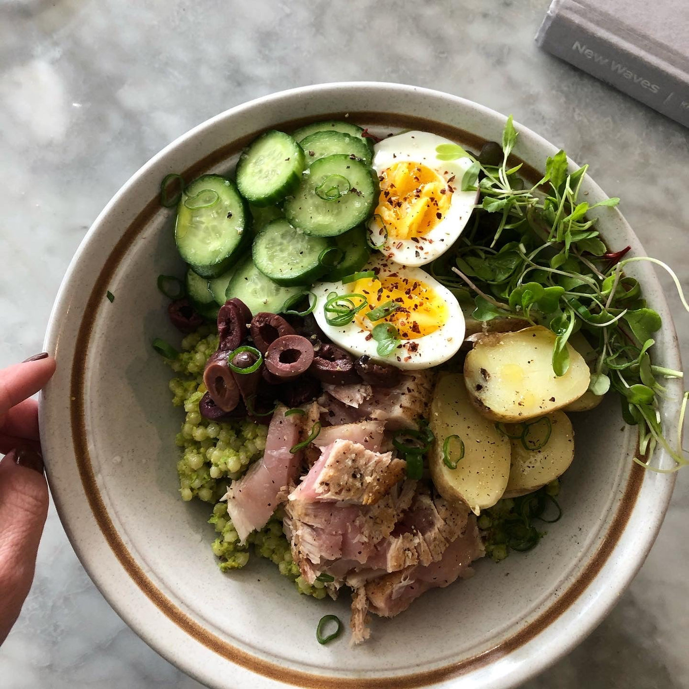 A Niçoise salad in a bowl. The salad contains grains, greens, potatoes, eggs, cucumbers, olives, and tuna.
