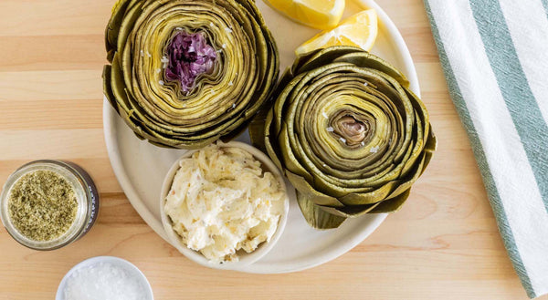 An artichoke, cut in half, with compound butter and lemon wedges.