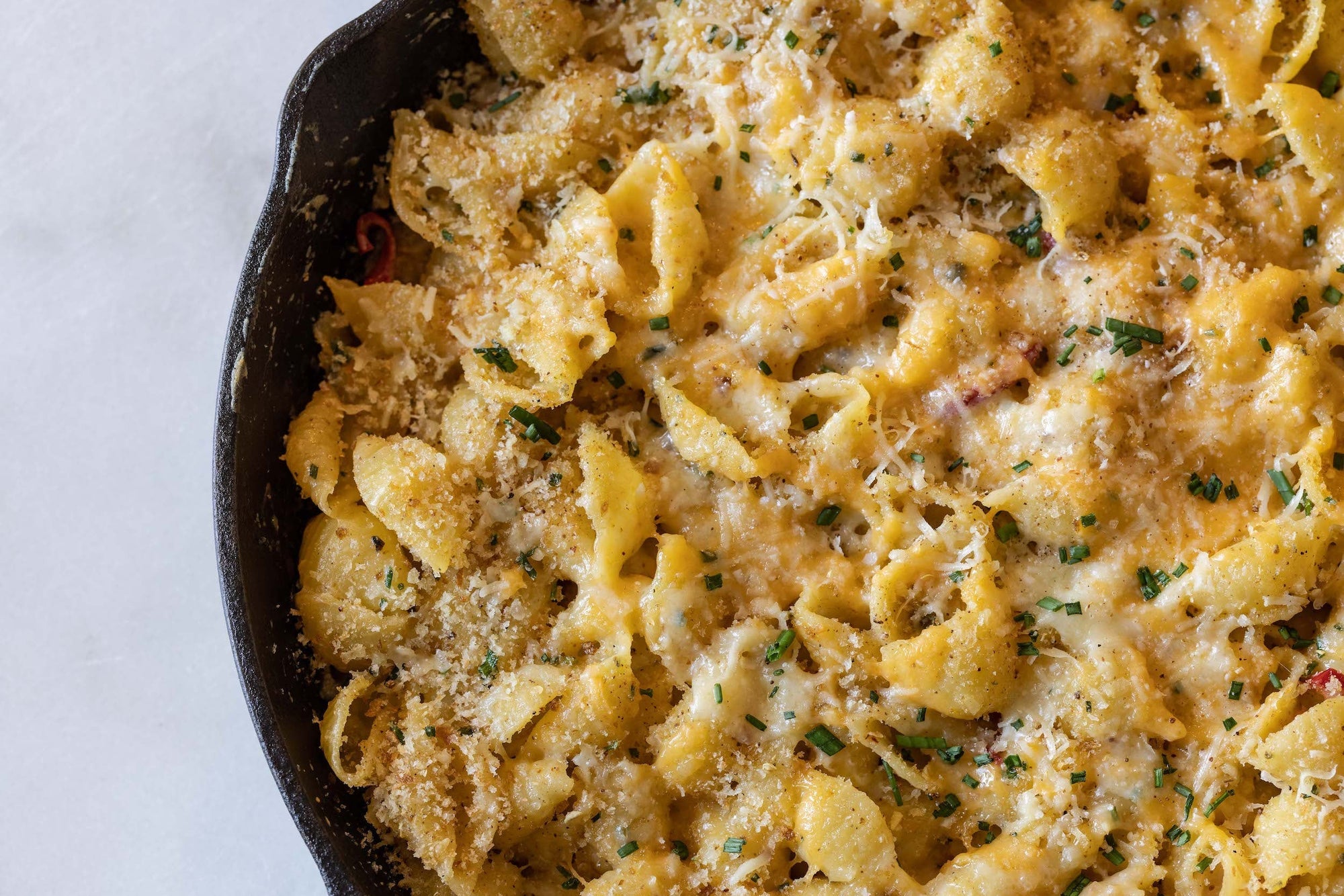 A cast-iron skillet is filled with macaroni and cheese, bread crumbs, melted cheese, and chopped chives.