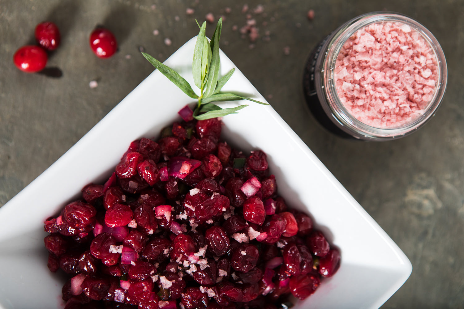 A square bowl of deep red cranberry salsa, next to a jar of pink pinot noir salt.