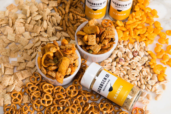 Various snack mix ingredients laid out on a table with jars of ramen seasoning, and two bowls with the snack mix and seasoning combined.