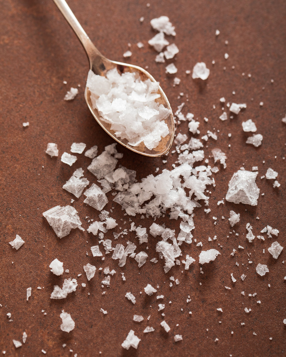 Large flakes of sea salt on a spoon, spilling out onto a brown countertop