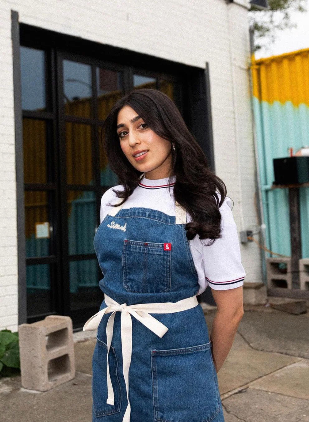 A woman, Samah Dada, stands with her hands behind her back. Her head is tiled to the side, she has a slight smile on her face, and she is wearing a denim apron that is tied in the front.