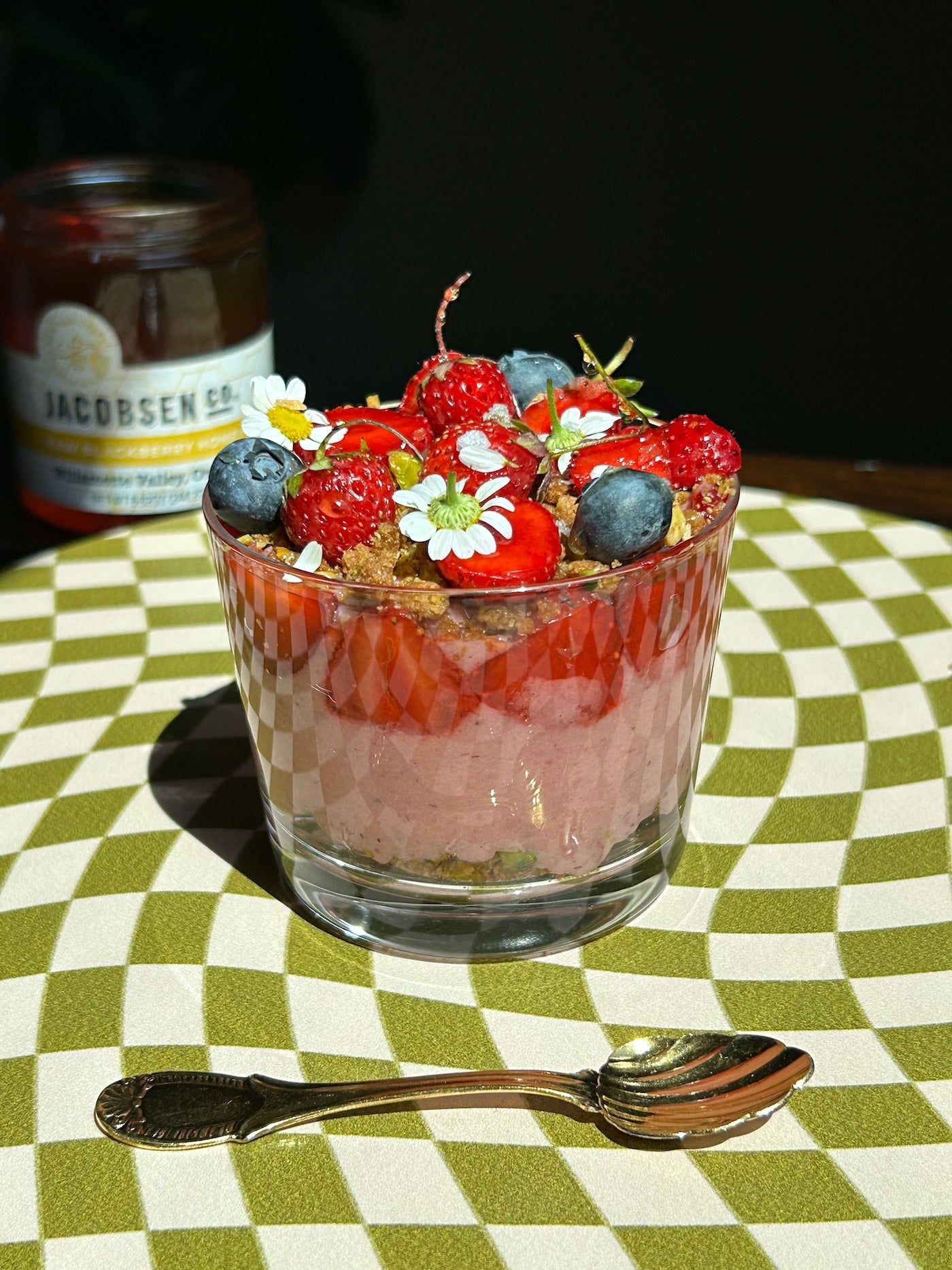Strawberry custard topped with whole strawberries, blueberries and edible flowers in a glass. The dessert is sitting on a green and white checkered tablecloth with a decorative spoon.