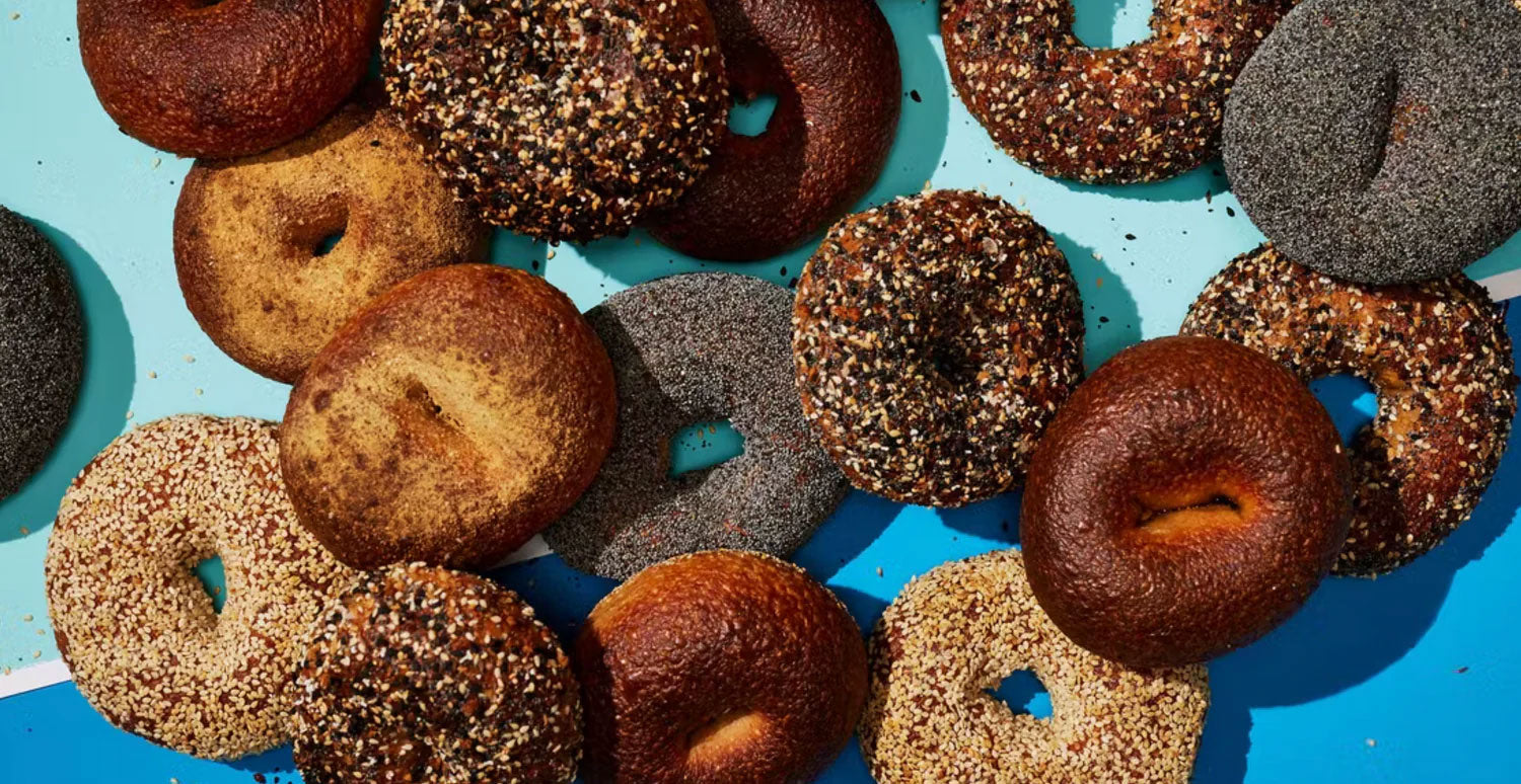 Sixteen bagels of various flavors are laid out on a blue backdrop.