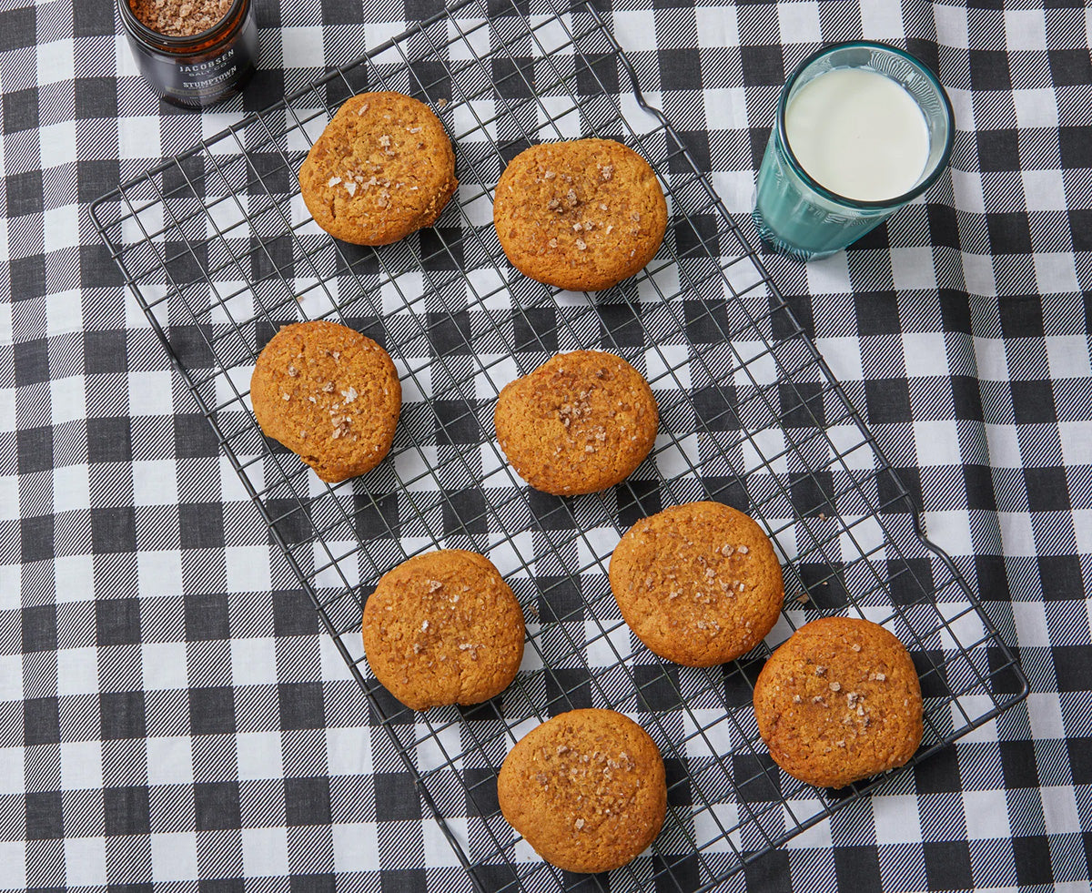 Ginger cookies cool on a black rack next to a glass of milk on top of a checkered tablecloth,