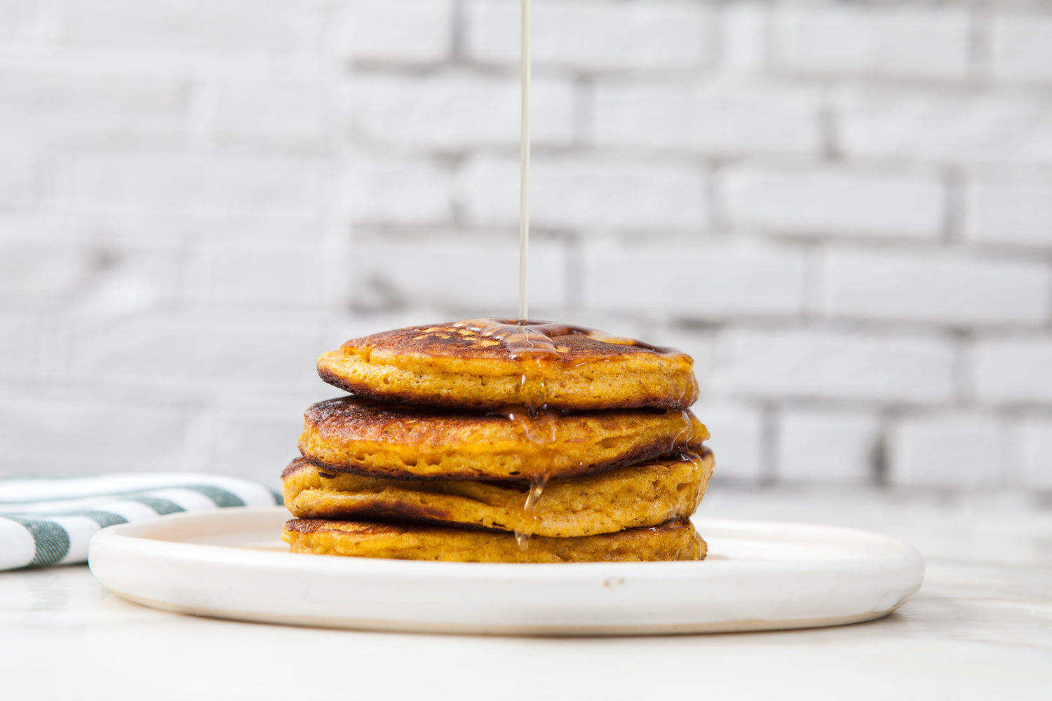 Pumpkin pancakes stacked on a plate, with syrup being poured over. 