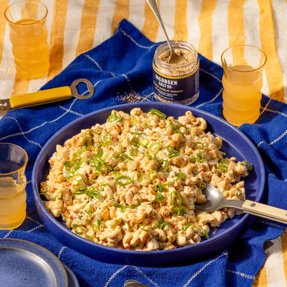 Macaroni topped with scallions, presented on blue plate on blue and yellow tablecloths, ingredients and drinks around the plate.