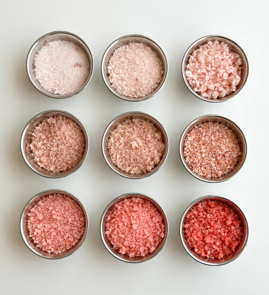 Nine circular tins of Koyo Berry Sea Salt. The top left tin is light pink, and the shades become darker in order until the bottom right, which is dark red.