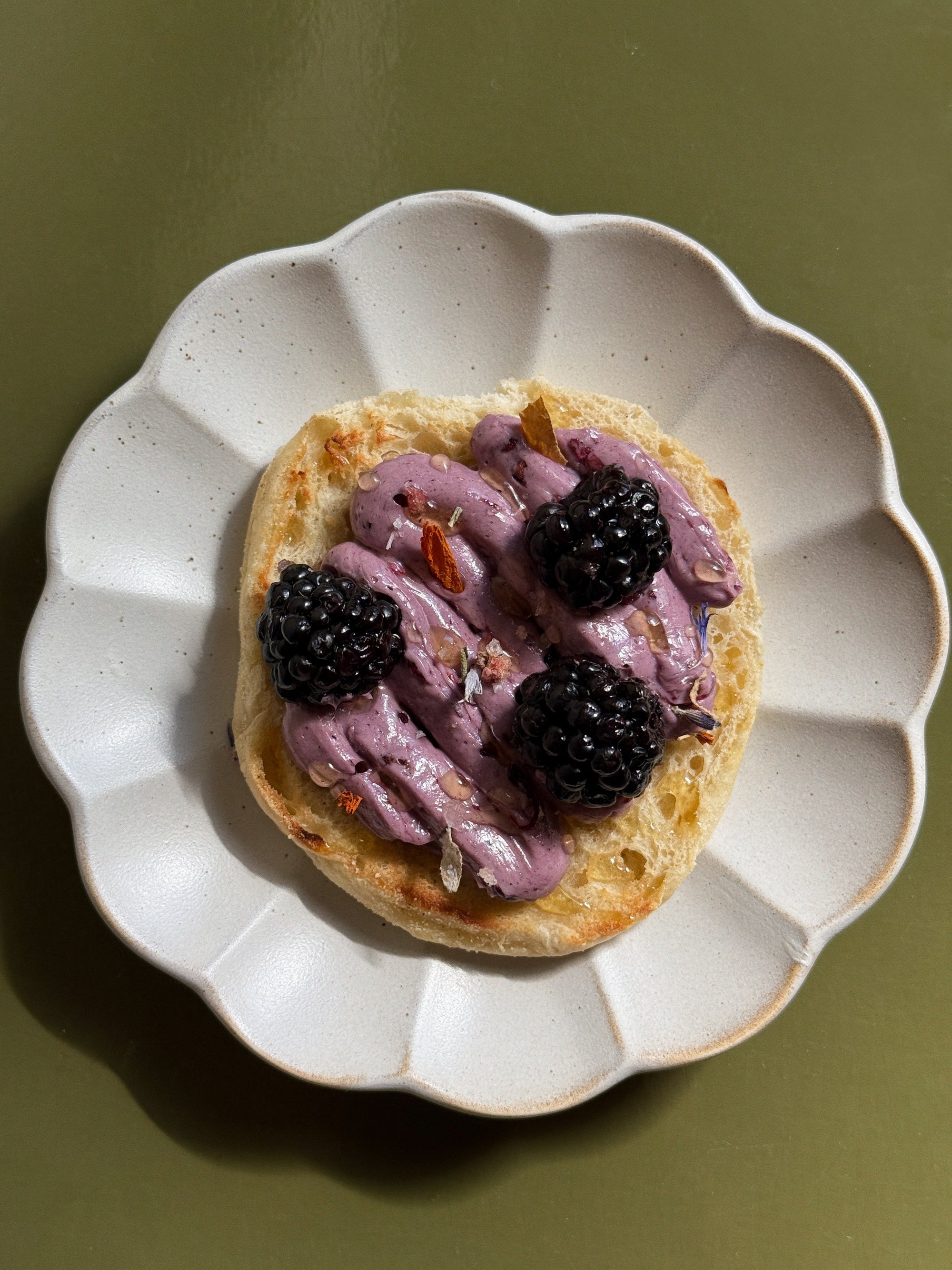 Plated english muffin, topped with purple whipped berry butter and blackberries on green backdrop.