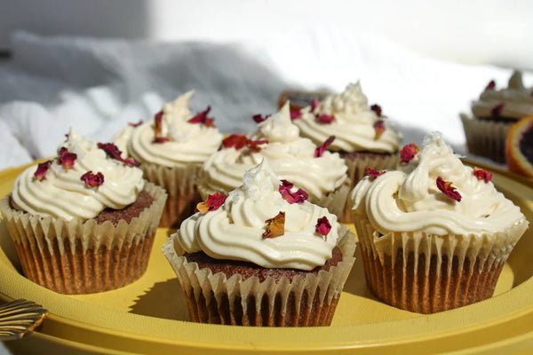 A plate of six cupcakes, decorated with frosting and rose petals. 