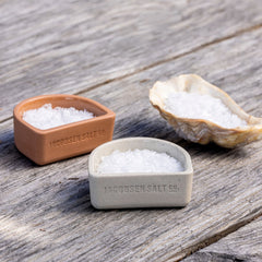 Ceramic salt cellars and a sea shell filled with salt on a wooden tabletop.