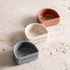 Three ceramic salt cellars, shaped like half circles in white, navy blue and canyon red with Jacobsen Salt Co. branding on the front.