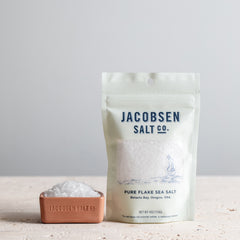 4oz bage of Jacobsen Pure Flake Sea Salt next to a canyon red salt cellar filled with sea salt. 