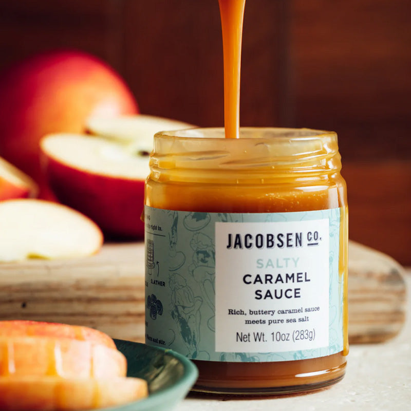 Jacobsen Caramel Sauce on a tabletop, with a spoon drizzling the sauce out of the jar. 