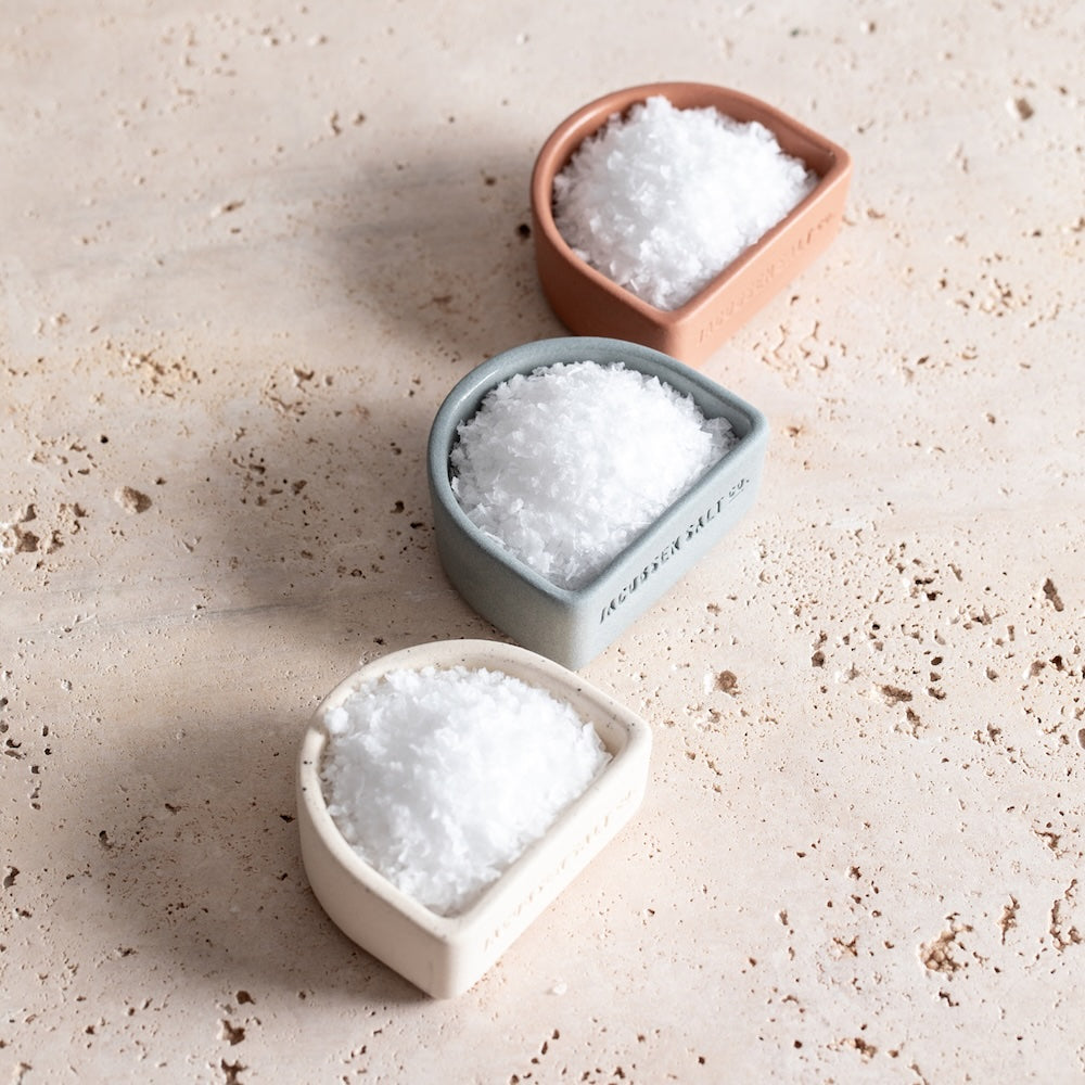 Three ceramic salt cellars, shaped like half circles in white, navy blue and canyon red. Each is filled with sea salt.