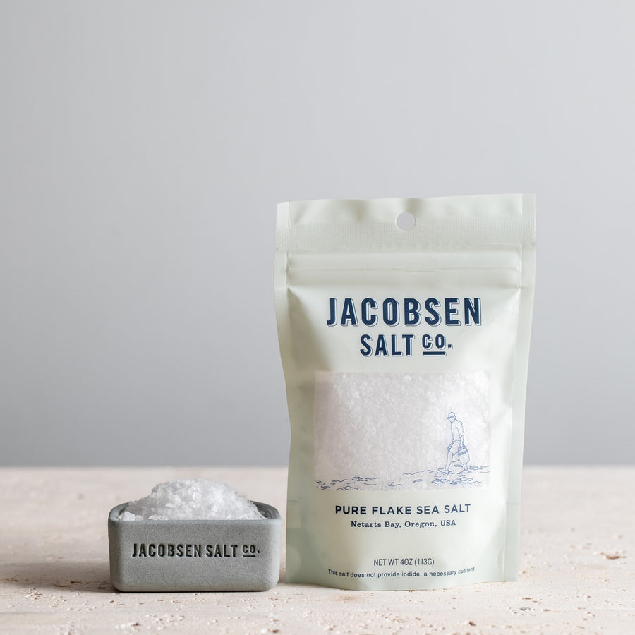 4oz bage of Jacobsen Pure Flake Sea Salt next to a cool grey salt cellar filled with sea salt. 