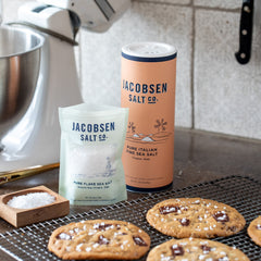 A bag of flake sea salt and a canister of fine Italian sea salt next to chocolate chip cookies 