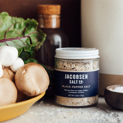 Jar of Jacobsen Black Pepper Salt on a countertop, next to a bowl of mushrooms and a salt cellar. 