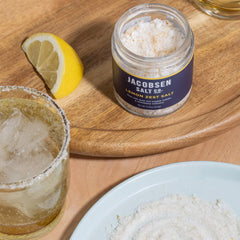 A jar of Jacobsen Lemon Zest Salt on a table with a beverage. The salt has been used to rim the drink.