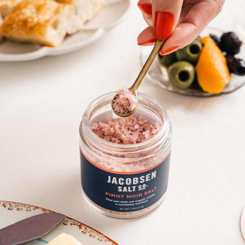 A bit of Pinot Noir Salt being scooped out of the jar by a tiny golden spoon, on an elegant tabletop.