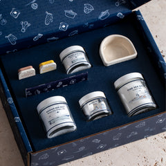 Various jars of sea salt, slide tins, and a white ceramic salt cellar in a navy blue gift box.