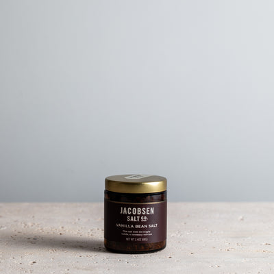 A  jar of Jacobsen Salt Co. Vanilla Bean Salt sits on a light stone surface against a soft gray background.