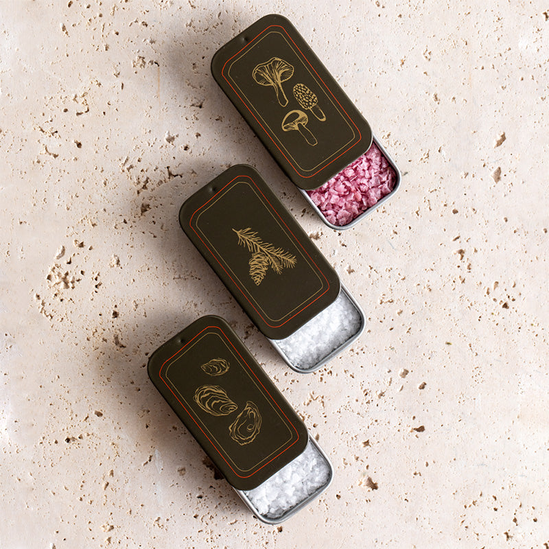 Three small rectangular dark brown metal tins decorated with gold botanical illustrations, arranged diagonally on a light beige textured stone surface. One tin is open, showing pink pinot noir salt inside, while the others show pure flake and pure kosher salt.