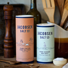 Two canisters of Jacobsen Salt (Kosher and Fine Italian), on a kitchen counter with wooden utensils in the background.