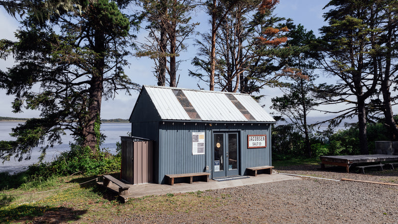 A small blue house structure next to the waters of Netarts Bay surrounded by sparse trees. The Jacobsen Salt Co gift shop.