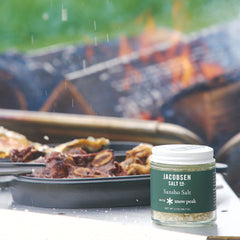 Jar of Jacobsen Salt Co. Sansho Salt on a table with food and grill in the background