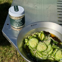 Cucumber salad with sesame seeds next to a jar of Jacobsen Salt Co. Sansho Salt on a metal surface outdoors.