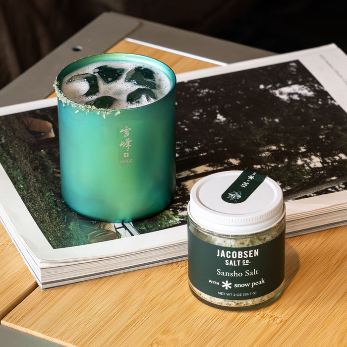 Green drink in a glass with a Jacobsen Salt Co. Sansho Salt container on a wooden surface.