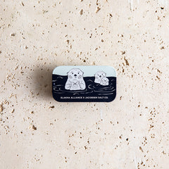 Small rectangular tin with illustrations of sea otters on a textured beige surface.