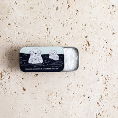 Open rectangular tin with illustrations of sea otters on a textured beige surface, with salt inside.