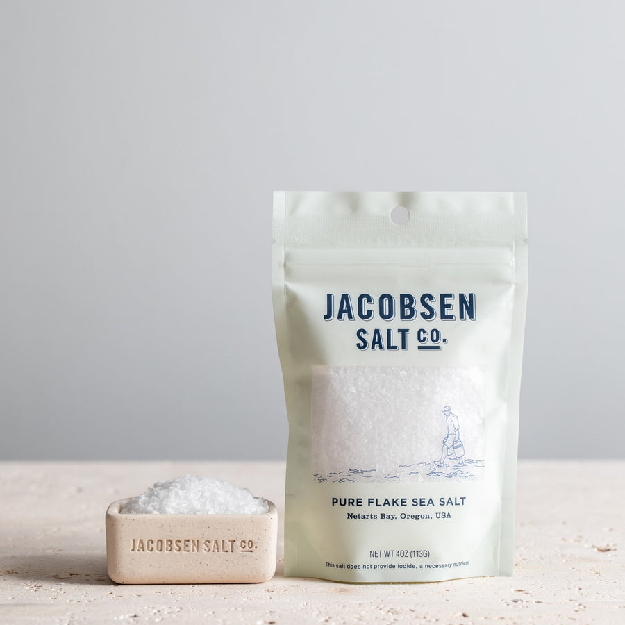 4oz bage of Jacobsen Pure Flake Sea Salt next to a speckled white salt cellar filled with sea salt. 