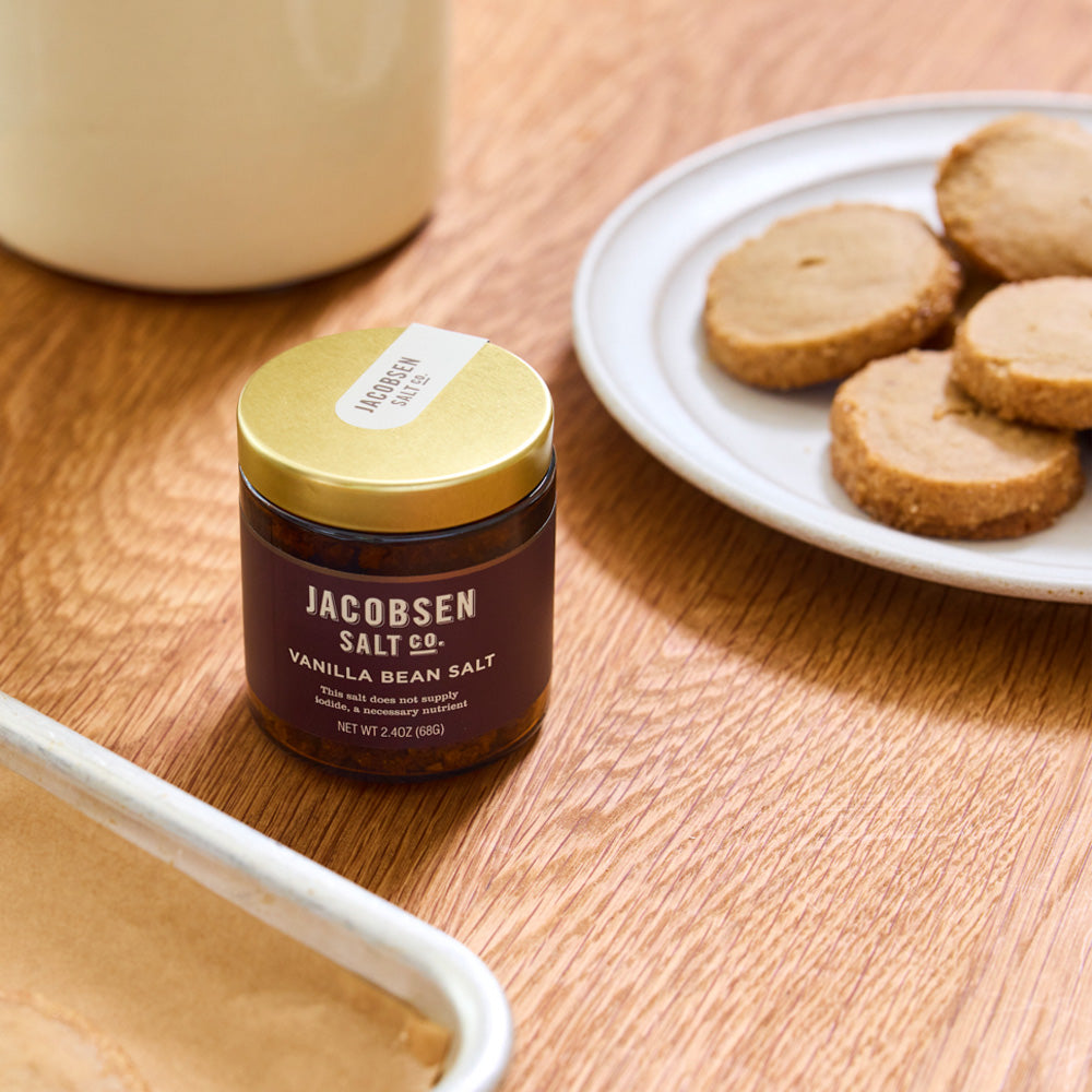A jar of Jacobsen Salt Co. vanilla bean salt sits on a wooden table beside a white plate of cookies.