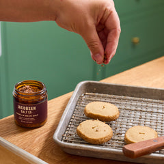 A hand sprinkles vanilla bean salt over three round cookies on a metal baking rack.